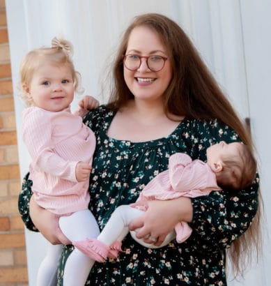 A woman holding her two daughters in front of a brick wall.